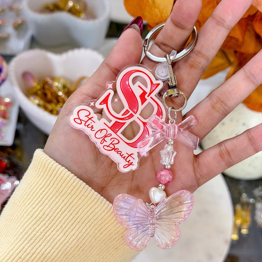 Hand holding a keychain with 'Stir Of Beauty' text and butterfly charm.