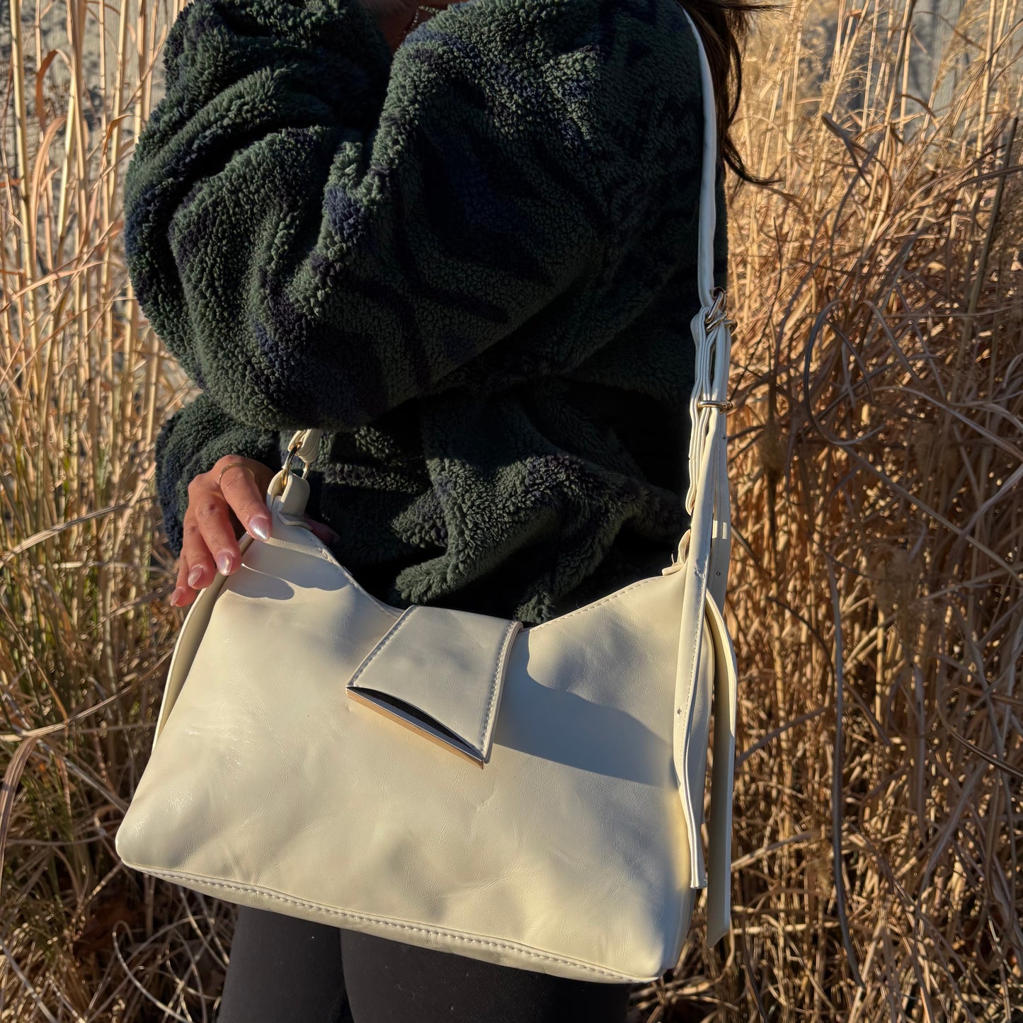 Ivory shoulder bag by Blaine, with a flap closure, held against tall grasses.