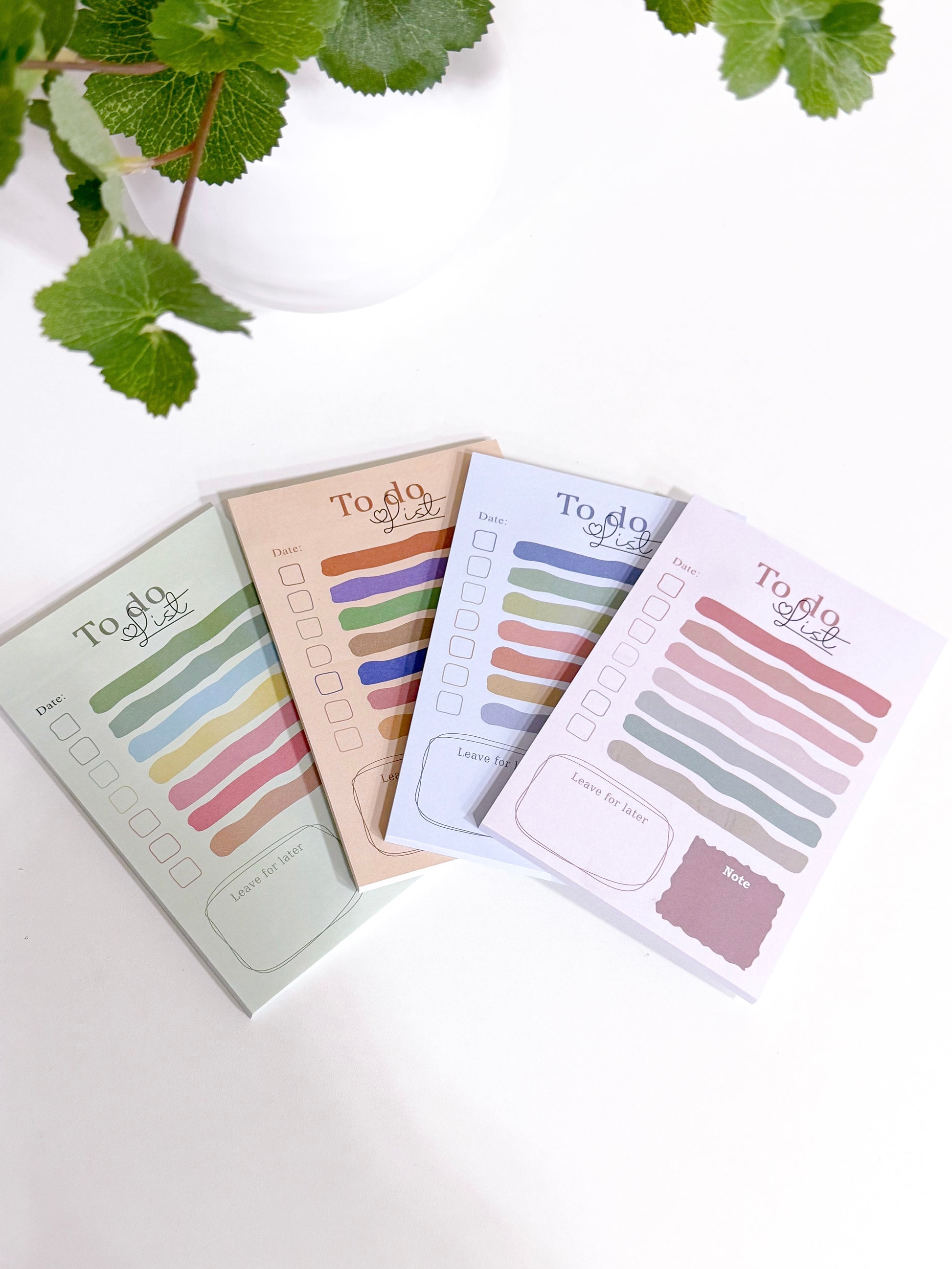 Four pastel To Do List notepads, each a different color, stacked on a white surface with green foliage.