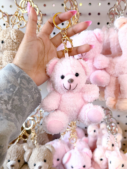 Pink plush Cuddle Bear Bag Charm with gold keyring, held by a hand against a background of other bear keychains.