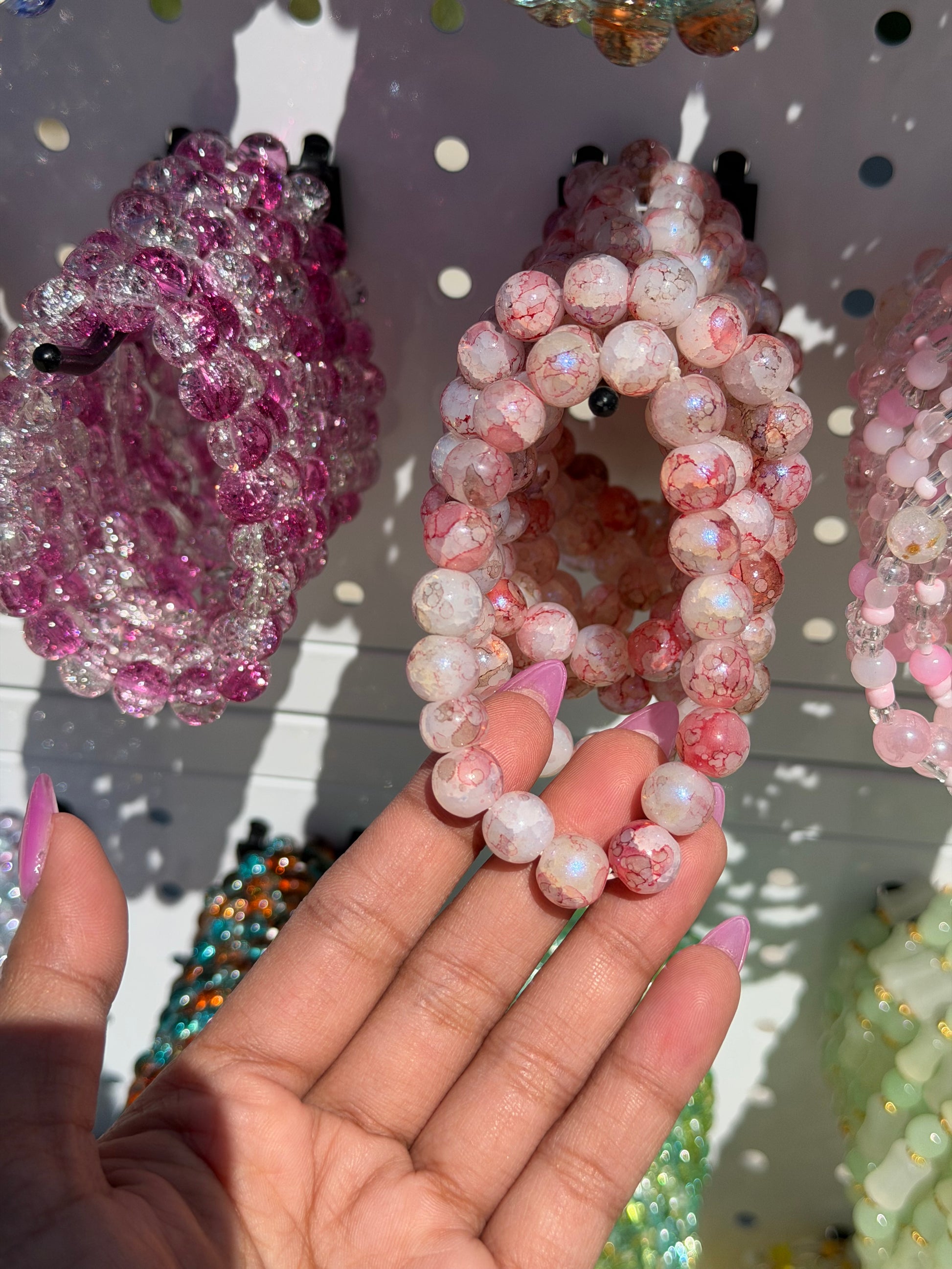 Pink beaded bracelets in two styles: one with crackled pink beads, the other with glittery pink and clear beads, held by a hand.