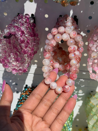 Pink beaded bracelets in two styles: one with crackled pink beads, the other with glittery pink and clear beads, held by a hand.