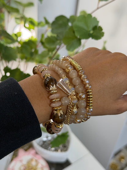 Layered beaded charm bracelets in light brown, dark brown, and gold with faceted and smooth beads, gold rings, and a clear pointed charm, worn on a wrist.