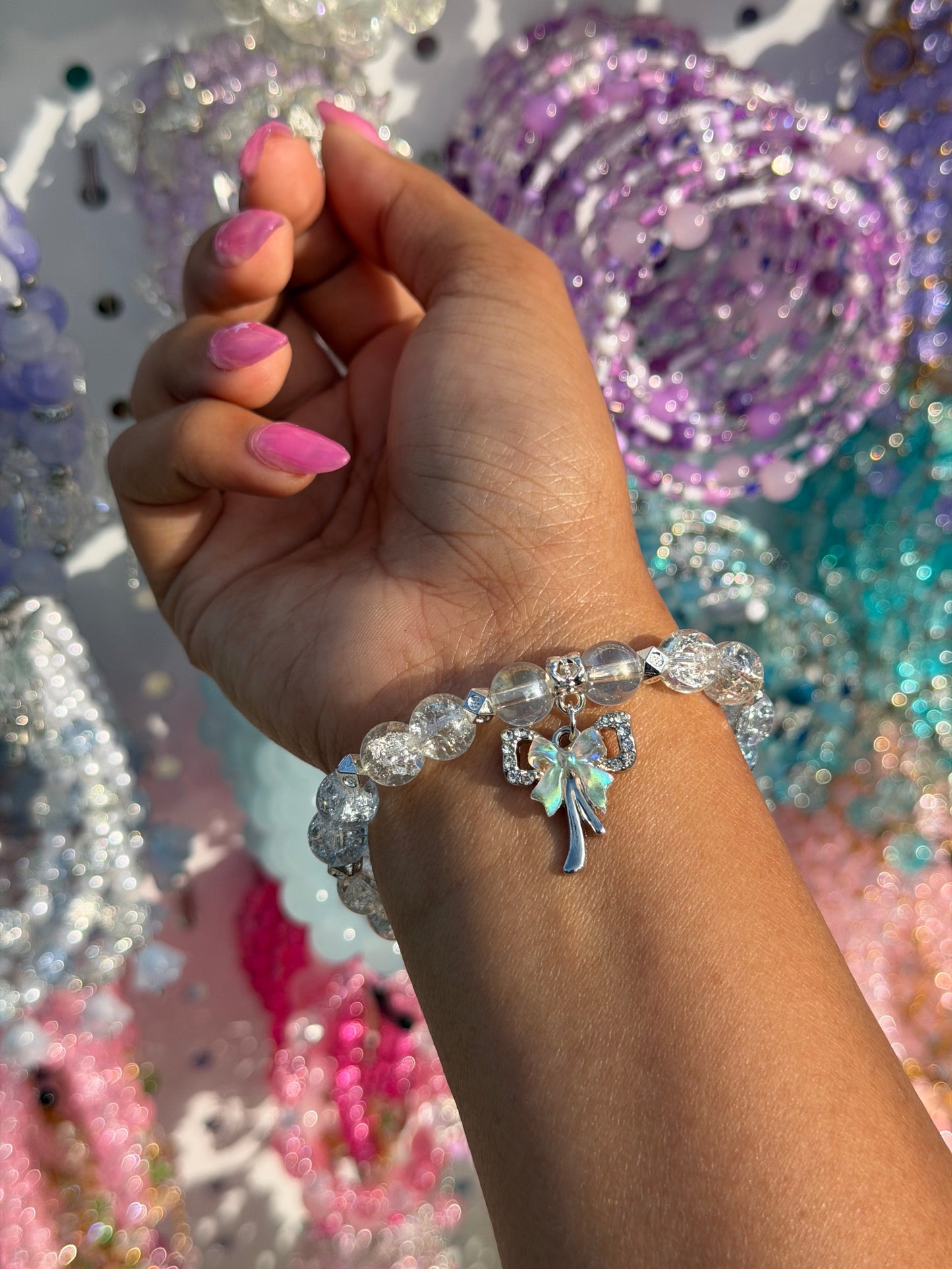 Beaded bracelet with clear cracked beads and an iridescent bow charm on a wrist with pink nails.