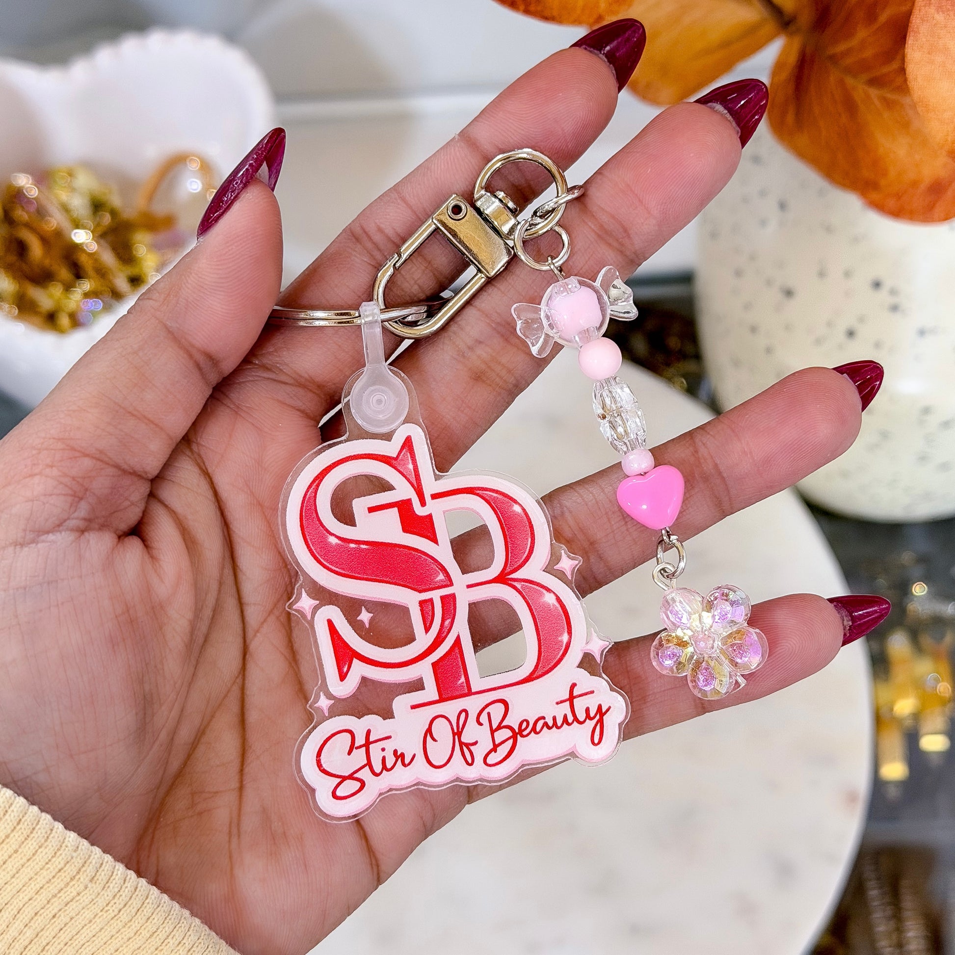 Hand holding a keychain with 'Stir Of Beauty' logo and decorative elements.
