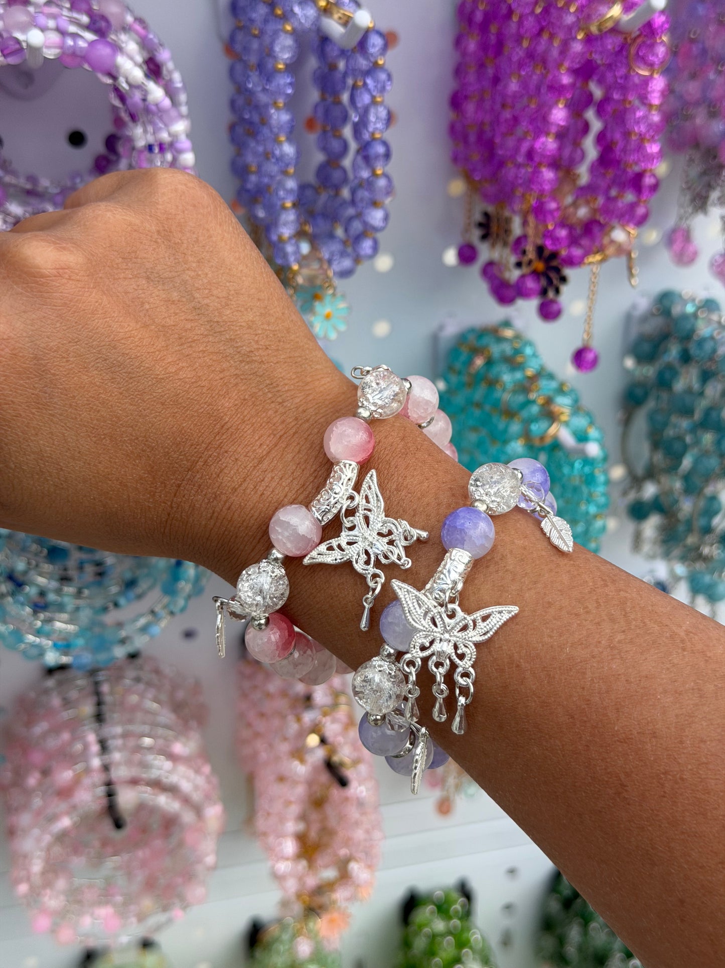 Two silver butterfly charm beaded bracelets: one pink and clear, one purple and clear, on a forearm with blurred jewelry background.