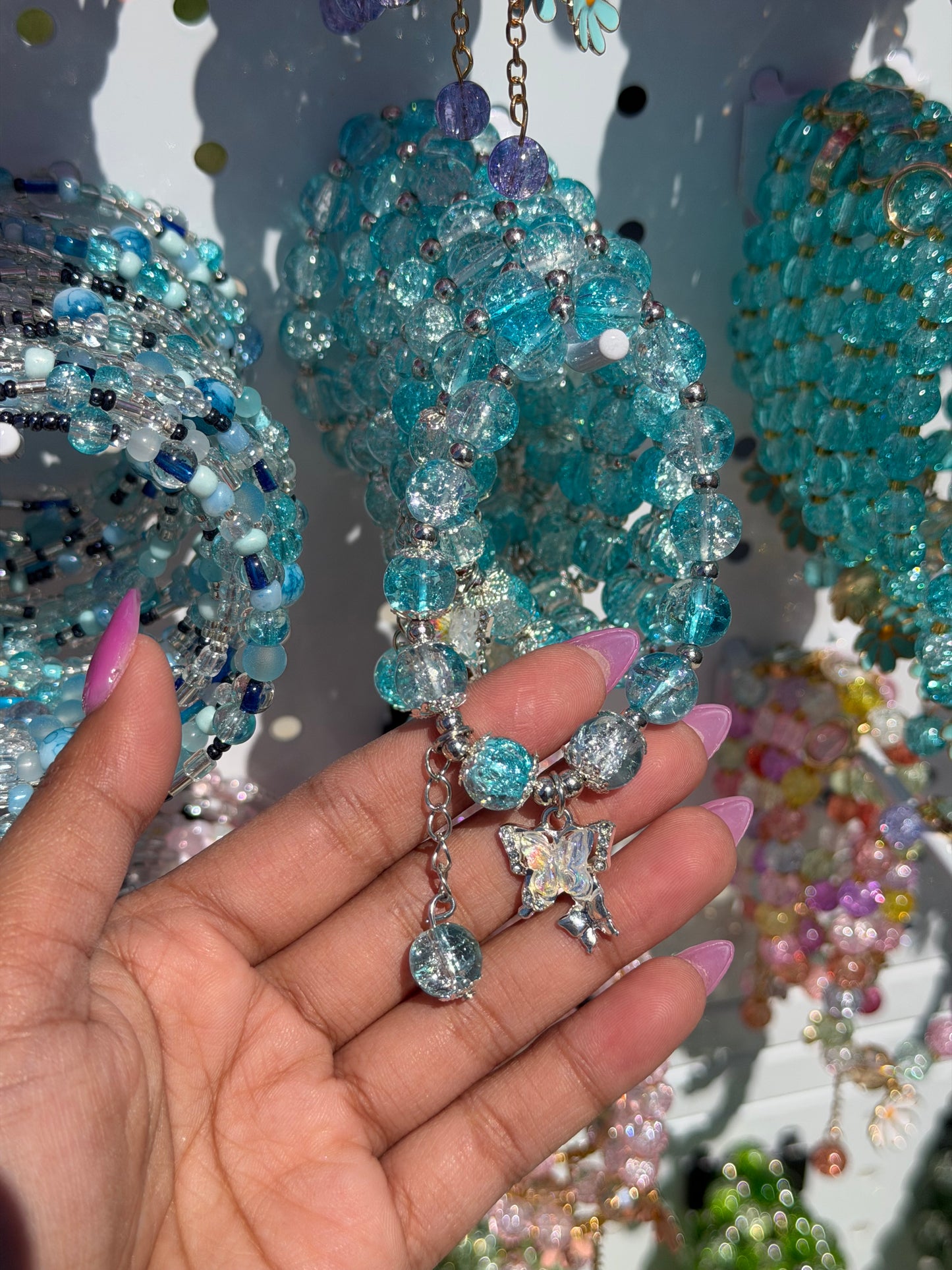 Blue beaded bracelet featuring clear and cracked blue beads with silver accents, a silver chain, and an iridescent butterfly charm, held by a hand with pink nails.