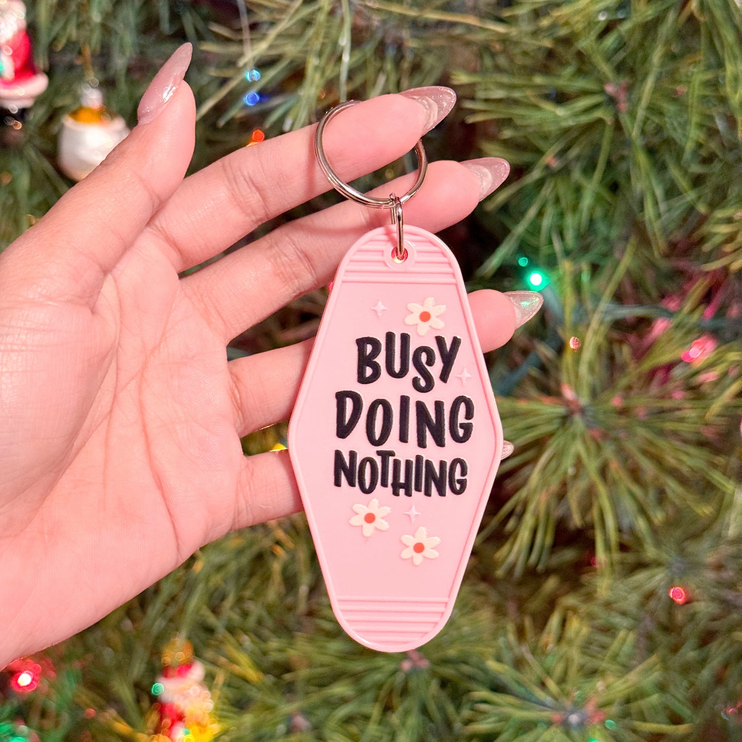 Pink motel keychain with "Busy Doing Nothing" text, decorated with small flowers.
