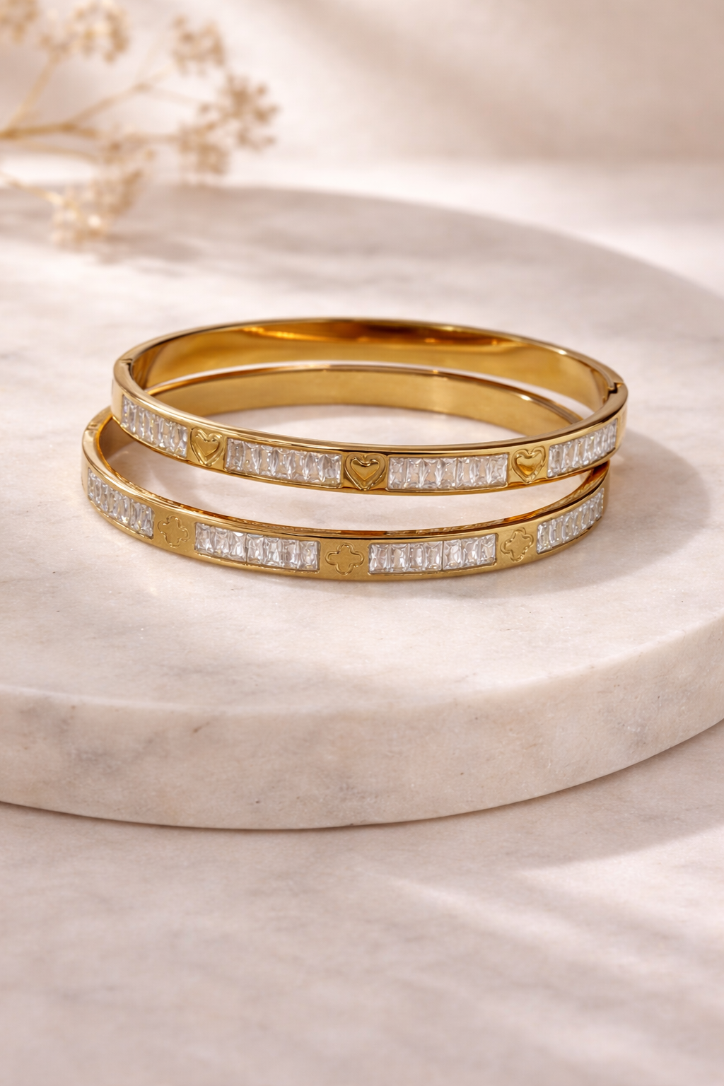 Two stacked gold bangles with baguette crystals; one has heart charms, the other clover charms, on marble.