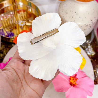 Back view of a white hibiscus-style flower hair clip held in hand, showing a silver metal barrette attached to the flower.
