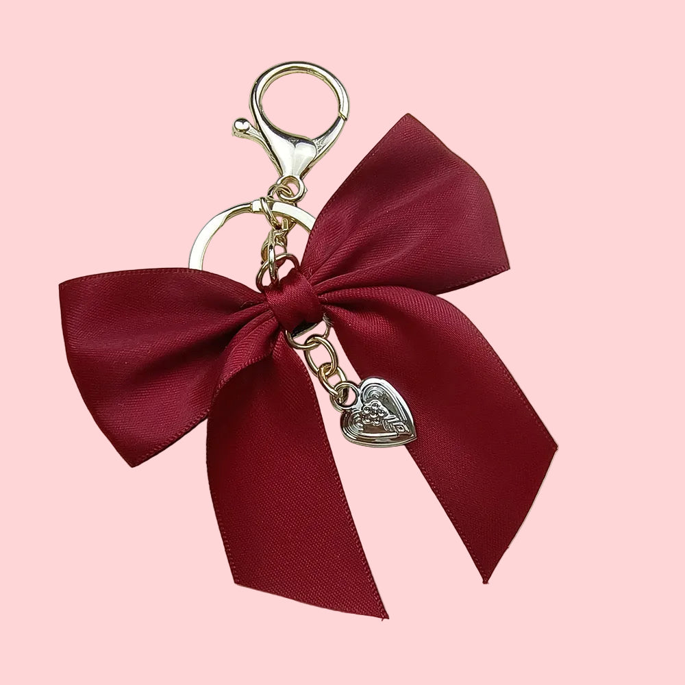 Dark red satin bow keychain with gold-colored hardware and a small silver heart charm on a pink background.