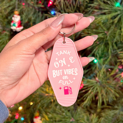 Pink motel keychain with the phrase, "Tank on E, But Vibes on Full" by Shop Amber Nicole held in a hand with long fingernails.