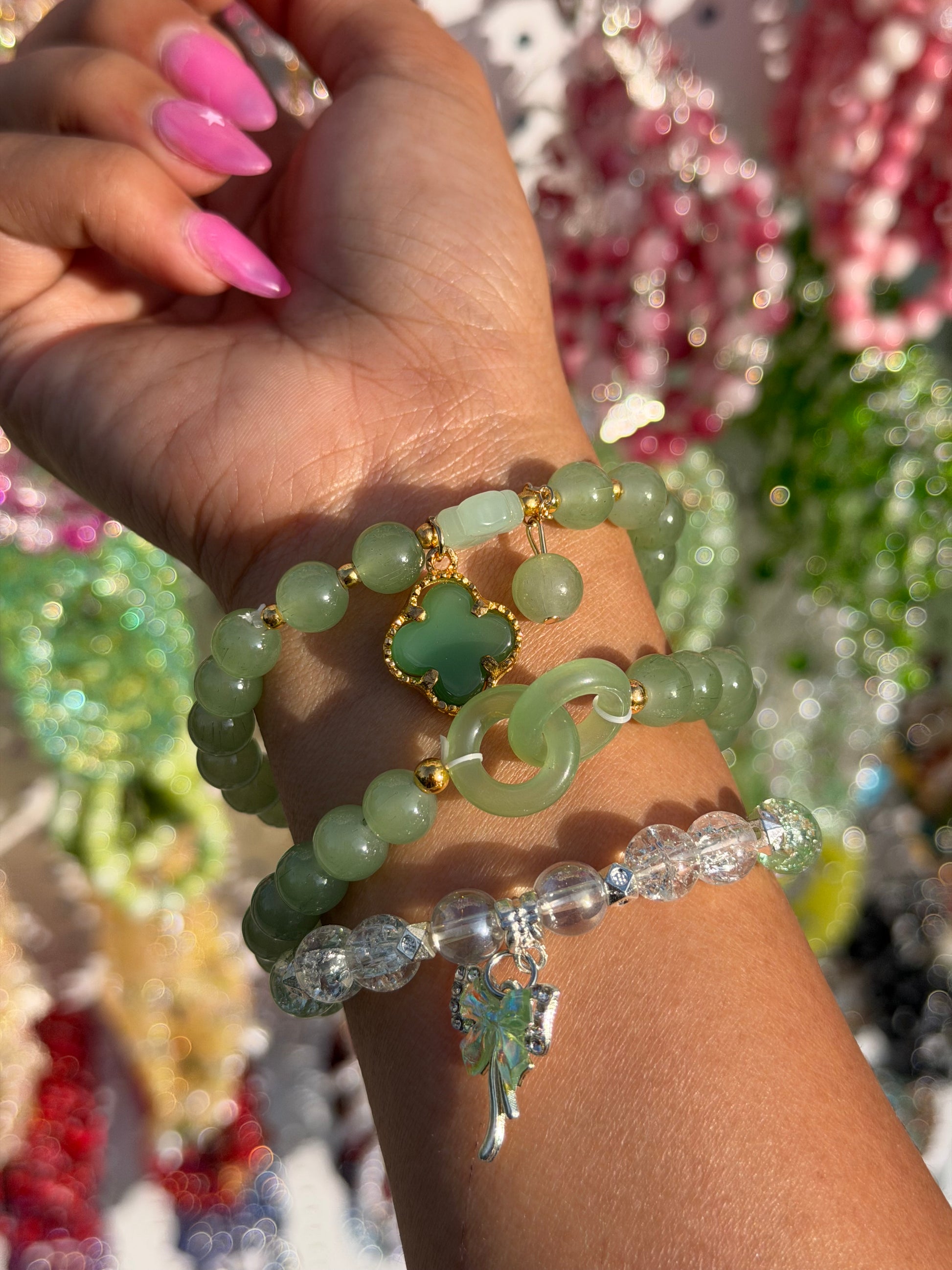 Green beaded lucky charm bracelets and a clear beaded bracelet worn on a hand with pink nails.