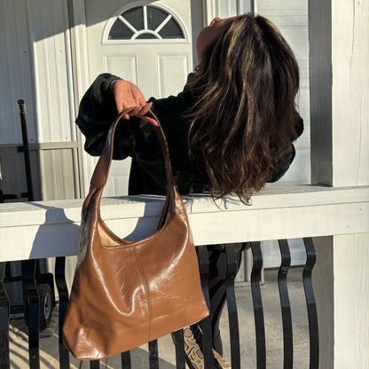 Brown tote bag with a hobo style shape, against a white railing and stylishly posed figure.