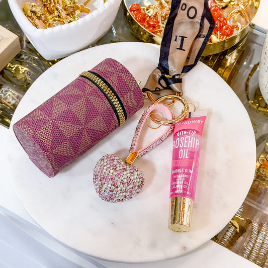 Pink geometric coin purse combo set with zippered coin pouch, rosehip oil lip gloss, and heart-shaped rhinestone keychain displayed on a white marble plate.