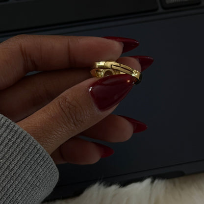Gold ring by Pavé held between two fingers, with dark red fingernails and a grey sweater cuff in view.