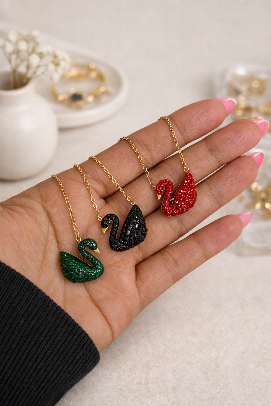 Three crystal swan necklaces in green, black, and red, with gold chains, held in a hand.