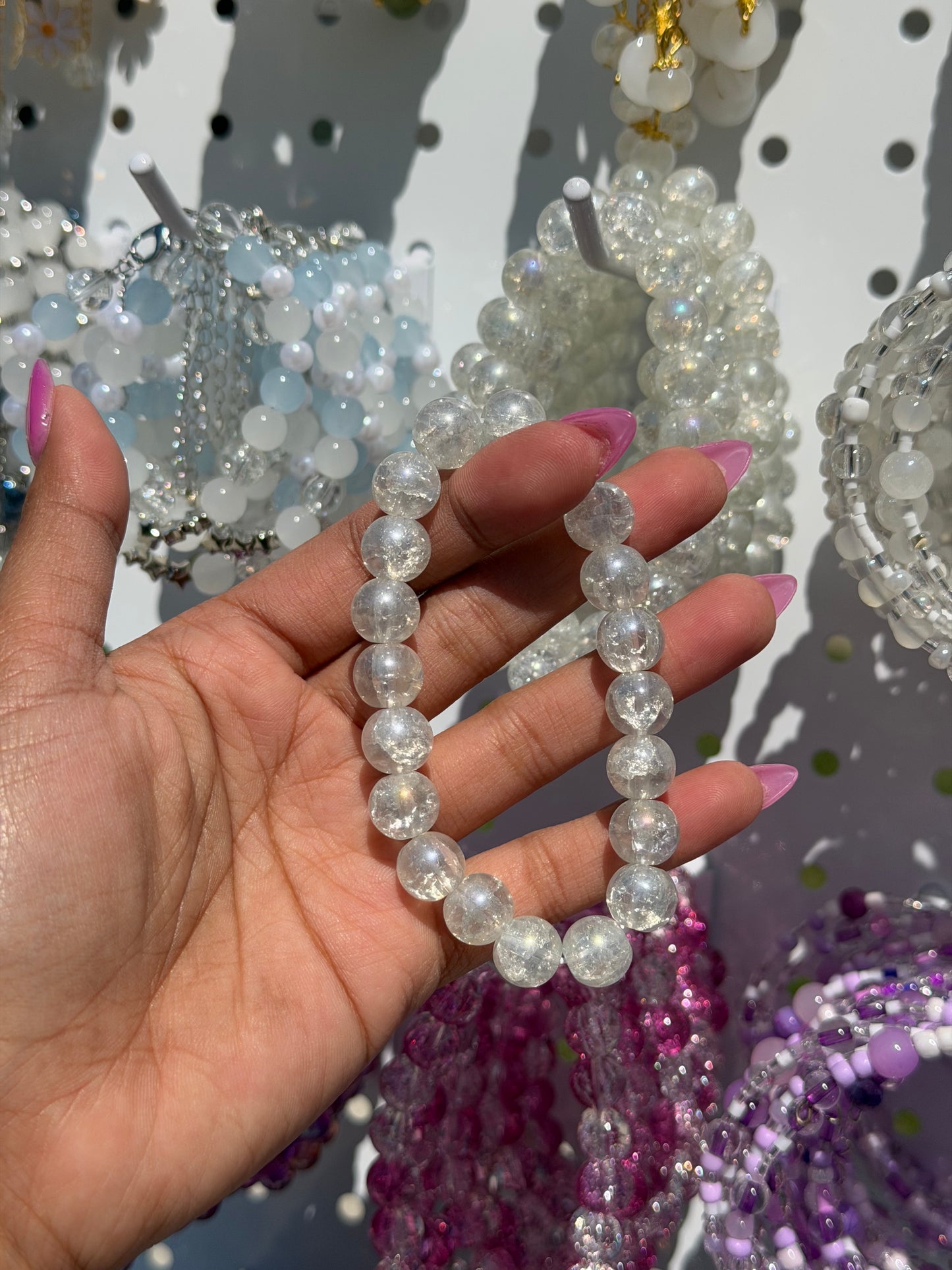White beaded bracelet with cracked clear beads, held by a hand with pink nails, surrounded by other colorful beaded bracelets.