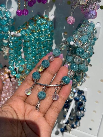 Blue beaded bracelet with cracked blue and clear iridescent beads, silver-tone bar, and dangling charms, held by a hand.