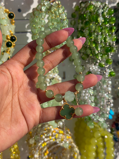 Green beaded bracelet featuring a light green flower and a dark green clover charm with gold accents, held by a hand.