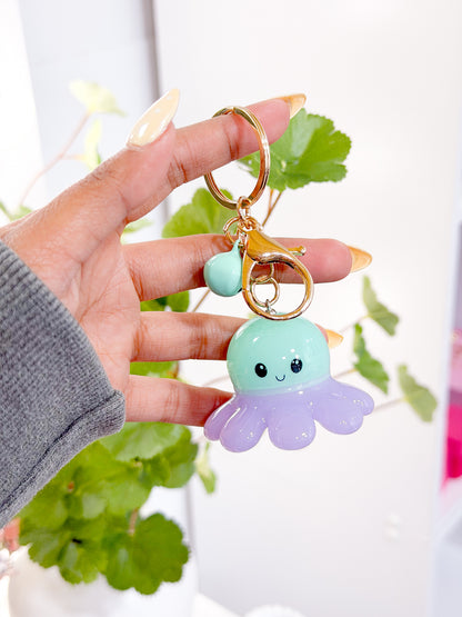 A two-tone mint green and lavender octopus charm with a cute face, on a gold-colored keychain with a small accent charm, held by a hand.