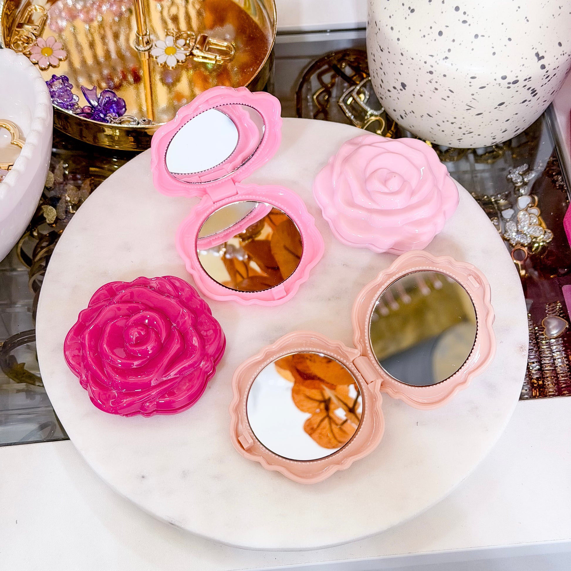 Rose-shaped compact mirrors in soft pink, blush, and hot pink displayed on a white marble tray