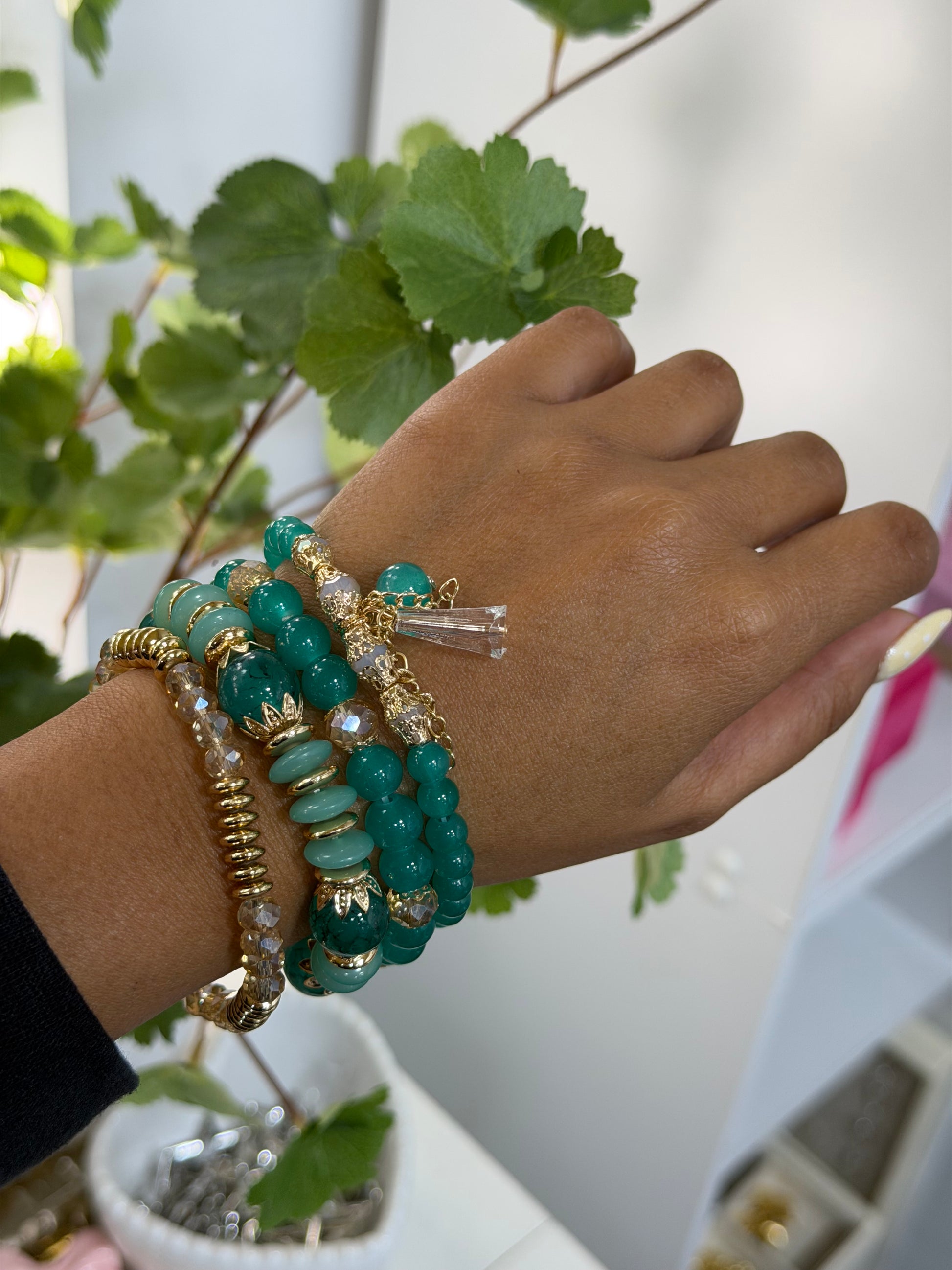 Teal and gold layered beaded charm bracelets with a clear rectangular charm, featuring varied bead textures, worn on a person's wrist.