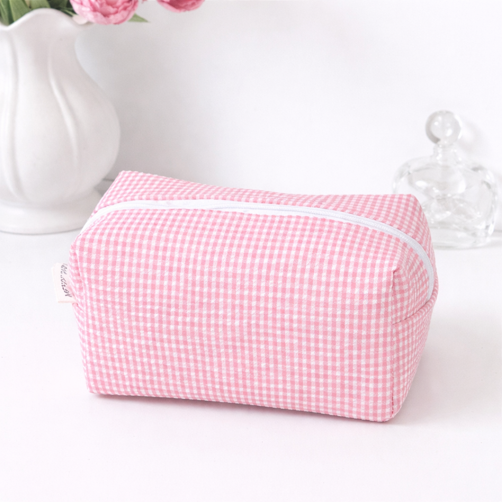 Pastel pink gingham makeup bag with a white zipper and a small side label, displayed with a vase and glass bottle.