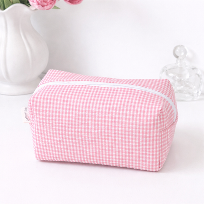 Pastel pink gingham makeup bag with a white zipper and a small side label, displayed with a vase and glass bottle.