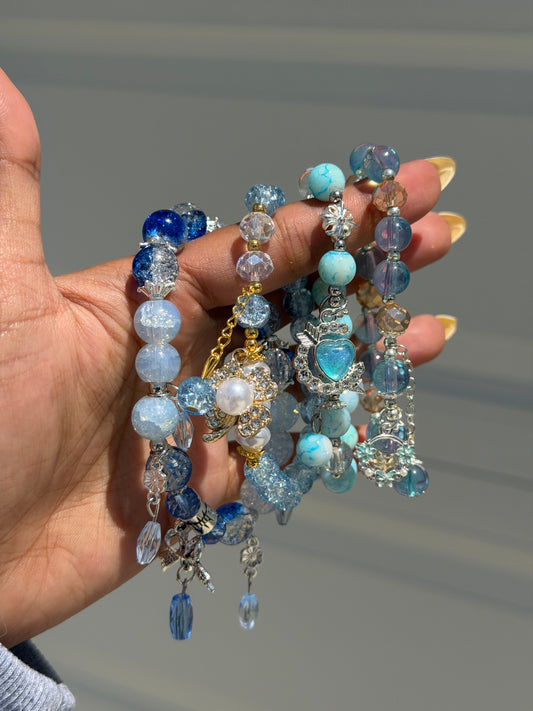 Blue charm beaded bracelet featuring various blue, clear, and glitter beads with silver-tone and gold-tone heart and butterfly charms, held in a hand.
