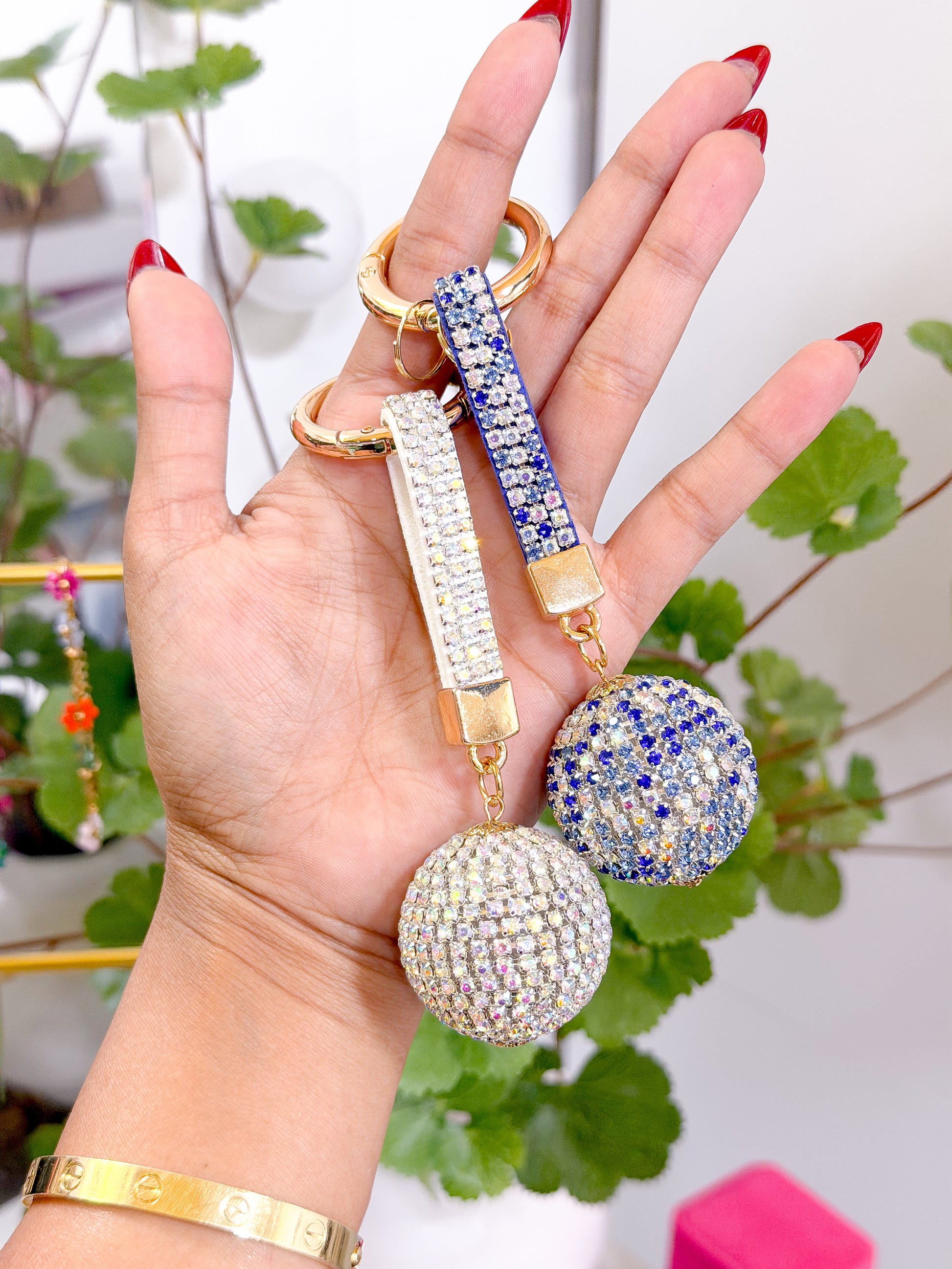 Shiny disco ball bag charms with crystal-encrusted spheres and rhinestone straps, held in hand with red nails, green foliage background