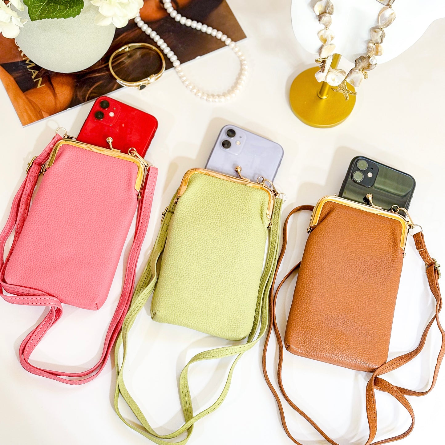 Three slim phone crossbody bags laid flat on a white surface in pink, light green, and tan, each with a gold clasp frame and long strap, shown holding smartphones partially inside.
