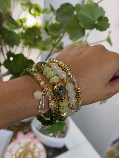 Layered green, white, and gold beaded charm bracelets with varied beads and a clear pendant, worn on a wrist, with blurred foliage.