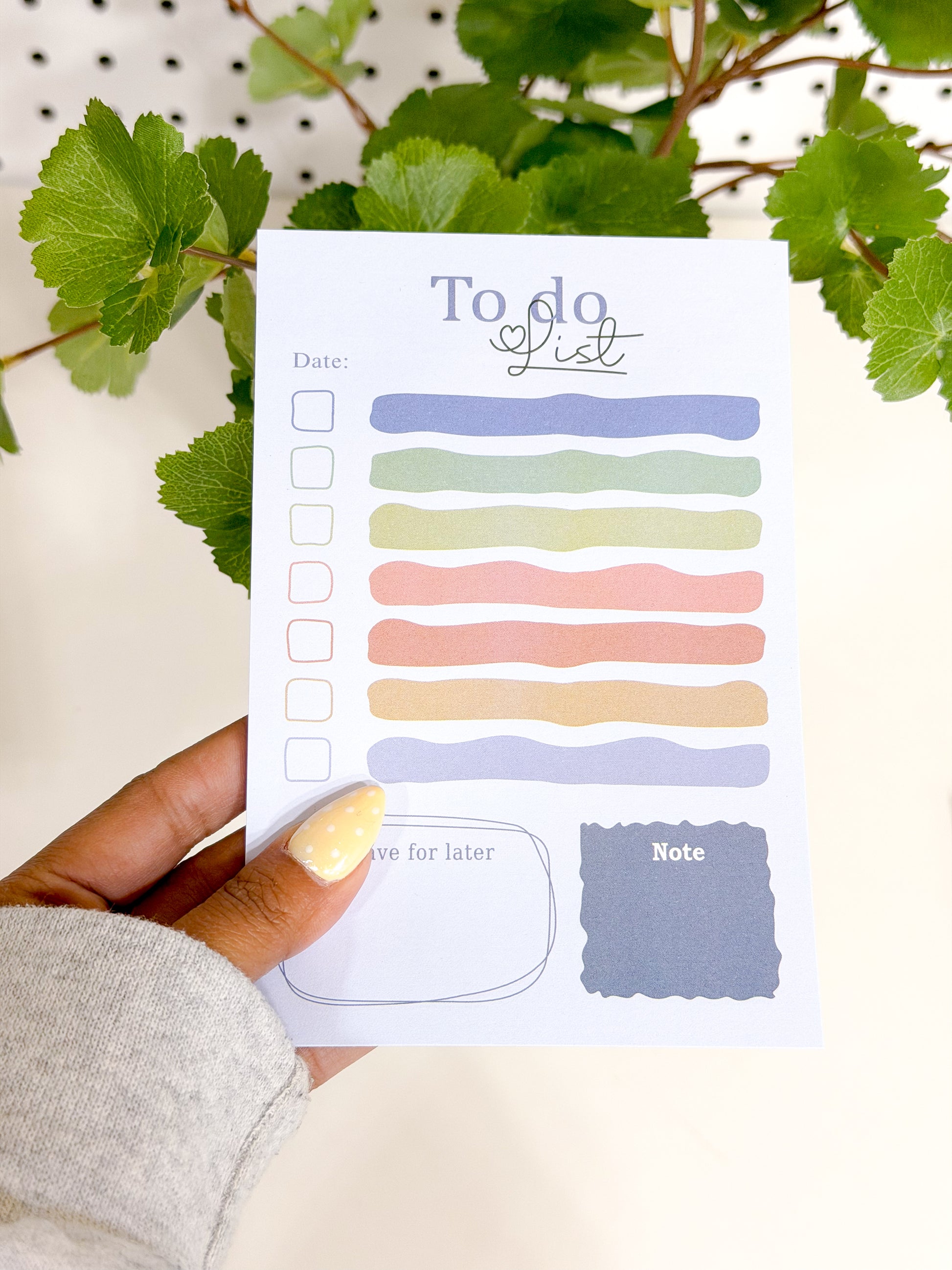 Pastel-colored To Do List Notepad with uneven lines and checkboxes, held by a hand with a yellow polka dot nail and a gray sleeve.