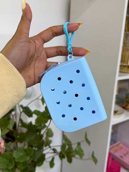Light blue silicone charm pouch with Crocs-like holes and a top zipper, featuring a matching clip, held in a hand.