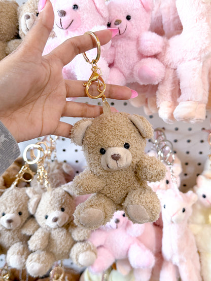 Tan fuzzy Cuddle Bear bag charm with a gold clip, held by a hand with pink nails, surrounded by other plush bears.