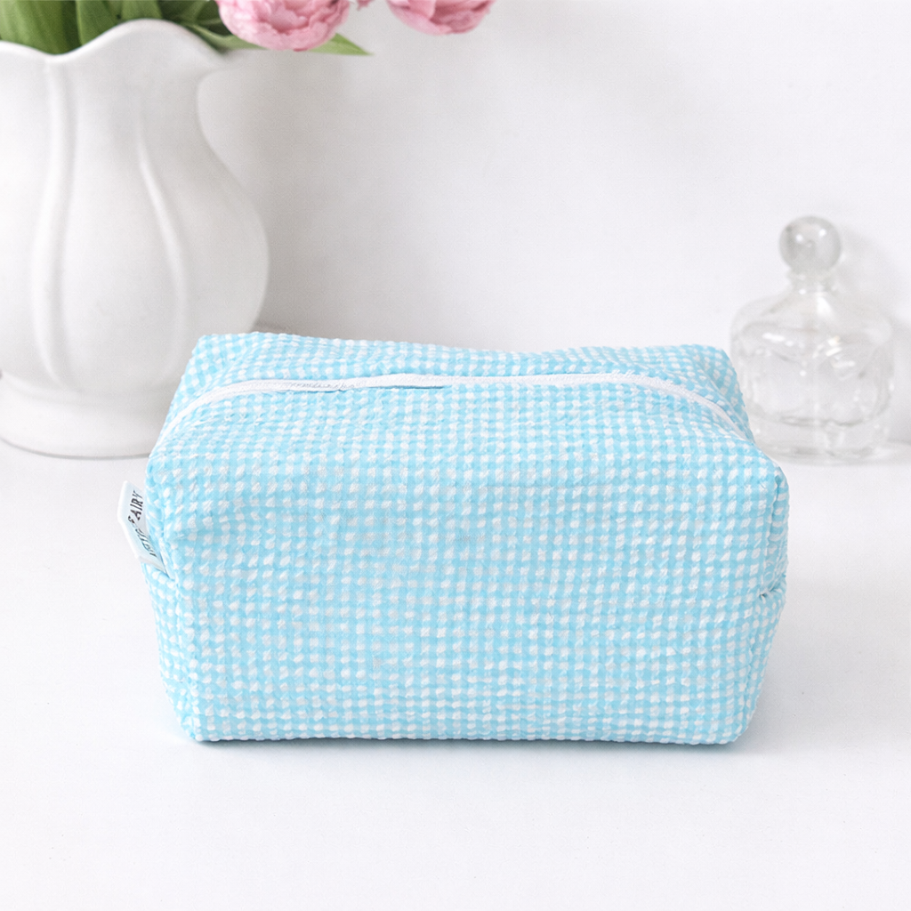 Pastel blue and white seersucker gingham makeup bag with a white zipper on a white surface.