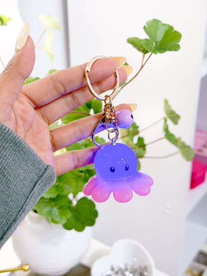 Glowing purple and pink octopus charm on a gold keychain, held by a hand with light nails.