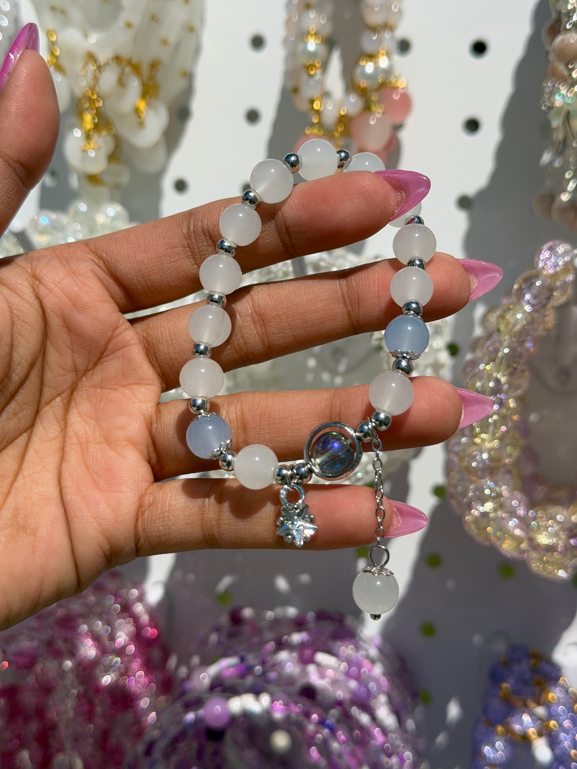 White beaded bracelet with light blue accents, silver spacers, and a star charm held by a hand with pink nails.