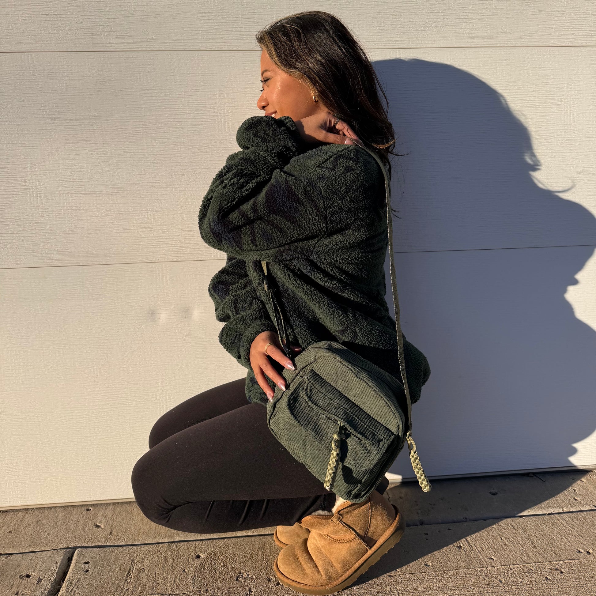 Green corduroy crossbody bag with a rope strap and front zipper, styled with a fleece jacket, leggings, and boots.