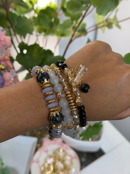 Layered beaded charm bracelets featuring black, lavender, and grey beads with gold accents and clear crystals, worn on a wrist.