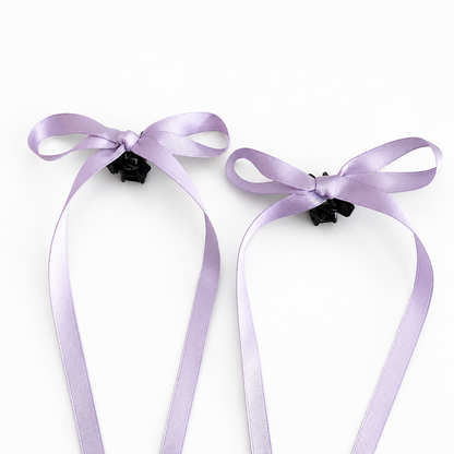 A set of two lilac satin bow mini hair clips with long trailing ribbons on a white background.
