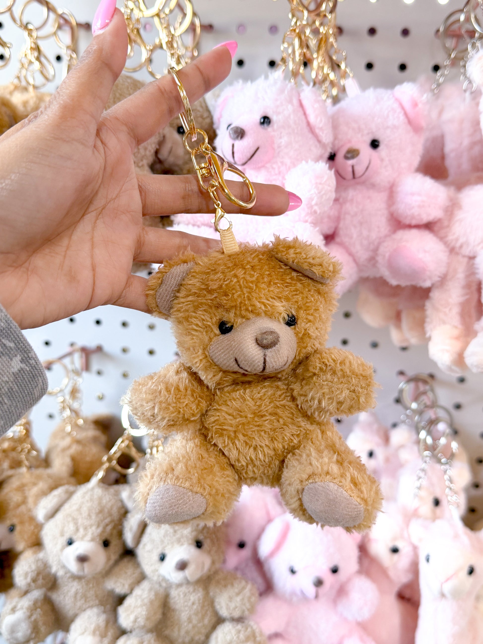 Brown fluffy Cuddle Bear Bag Charm with a gold keychain clip, held by a hand against blurred bear charms.