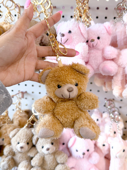 Brown fluffy Cuddle Bear Bag Charm with a gold keychain clip, held by a hand against blurred bear charms.