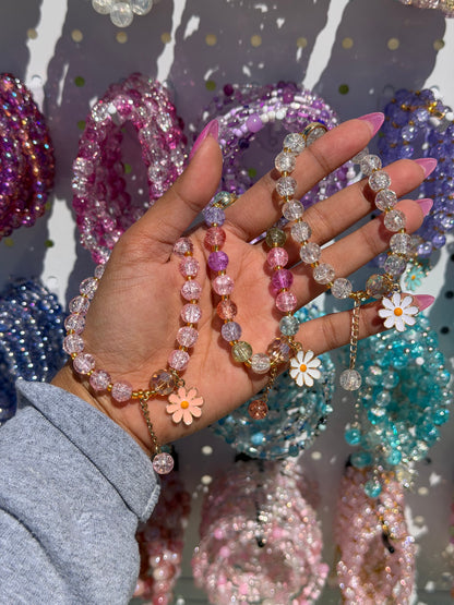 Multi-color and clear beaded bracelets with daisy charms, held by a hand in a grey sleeve against blurred bright jewelry.