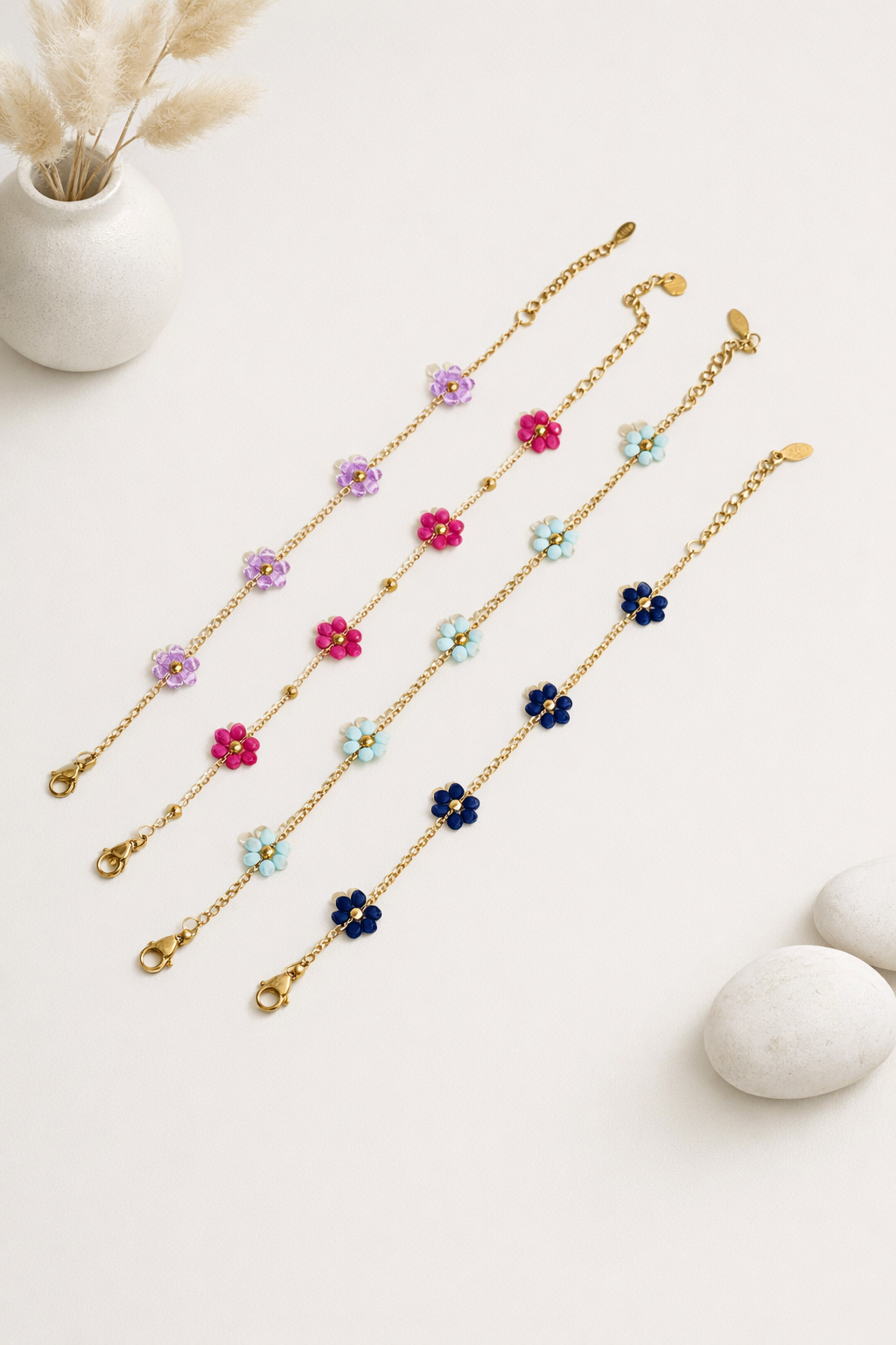 Four gold-toned Mini Daisy Bracelets with purple, fuchsia, light blue, and dark blue flower beads, laid diagonally on white.