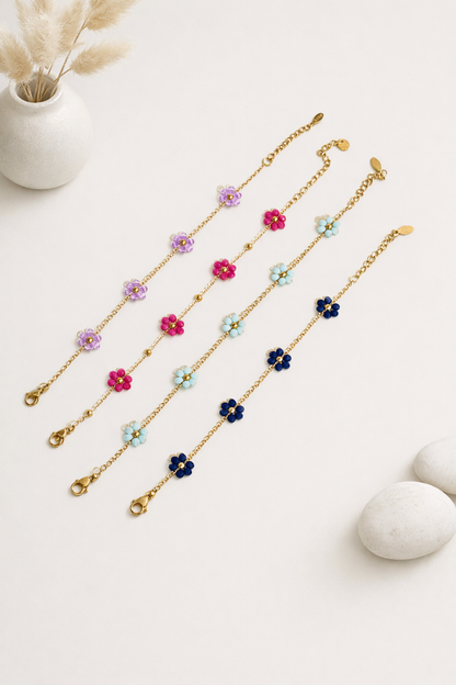 Four gold-toned Mini Daisy Bracelets with purple, fuchsia, light blue, and dark blue flower beads, laid diagonally on white.