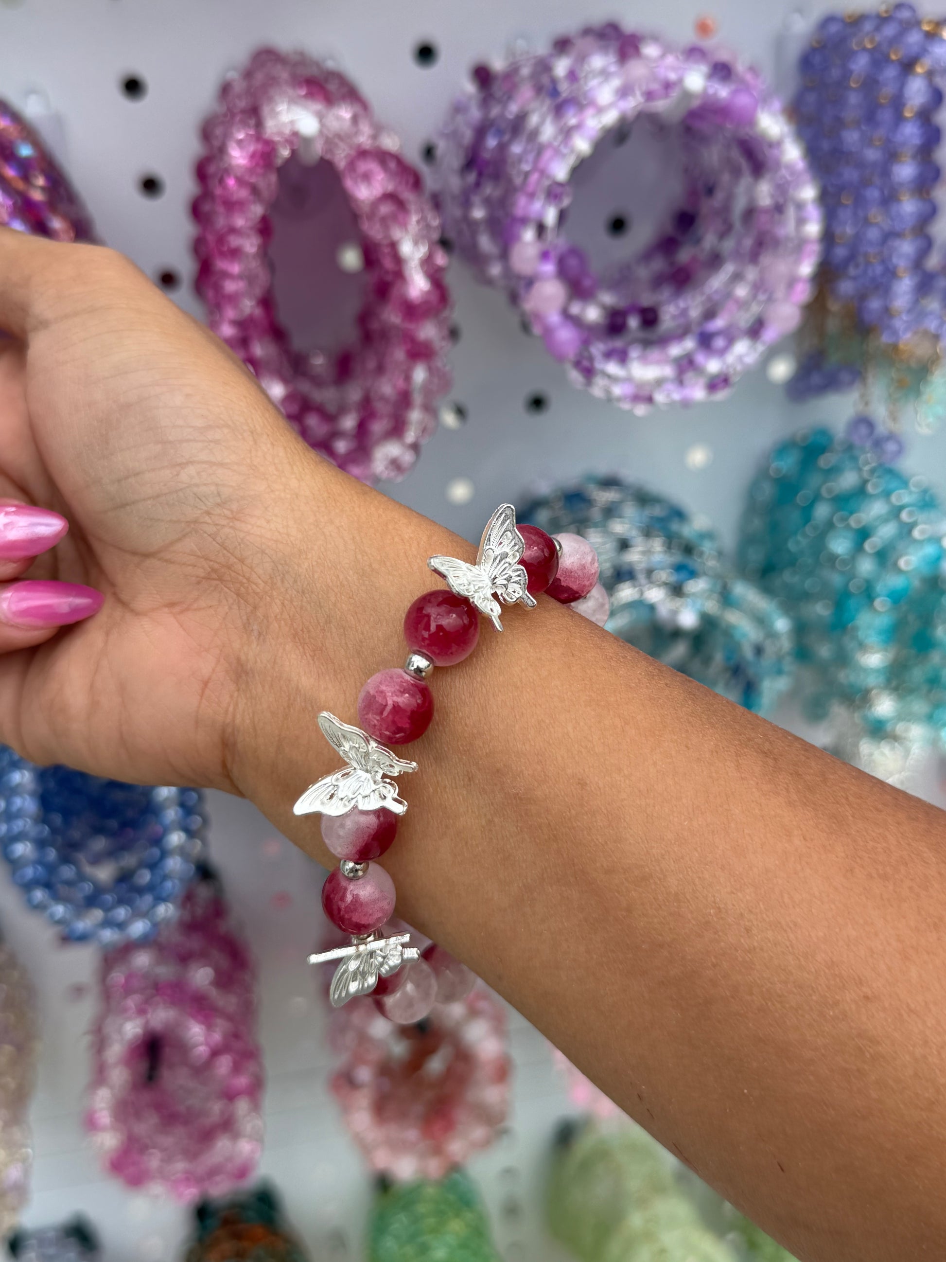 Red butterfly beaded bracelet with silver butterfly charms, worn on a wrist against blurred colorful bracelets.