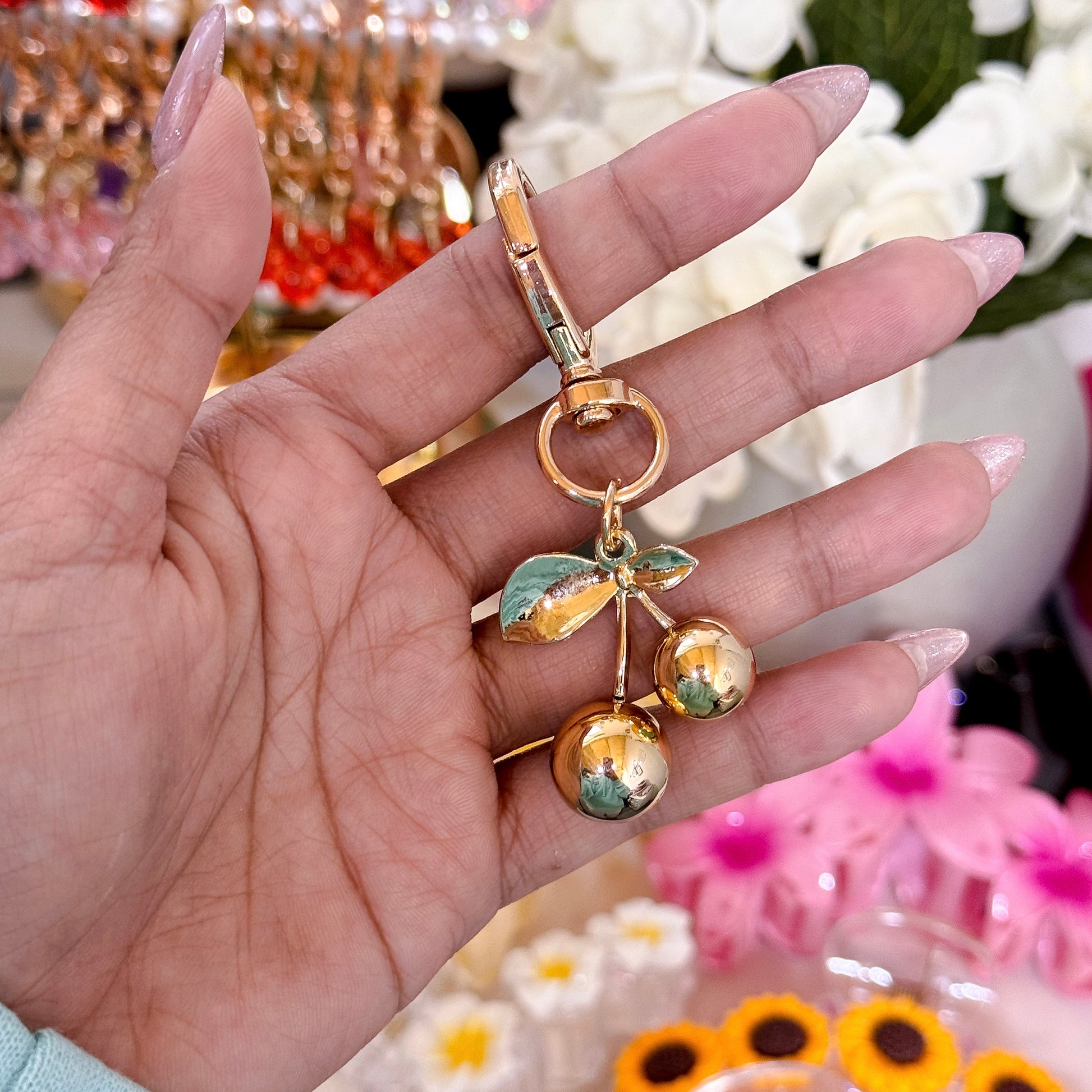 Gold heart-shaped clasp keychain with a dangling cherry charm featuring  gold cherries and gold leaves, held in a hand against a blurred floral background.