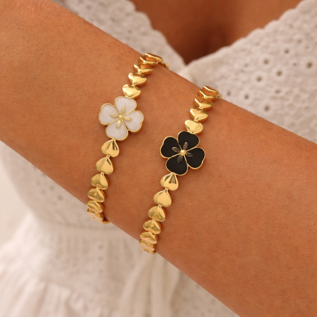 Gold-toned heart-link bangle bracelets with a white and a black flower charm, worn on a woman's arm.